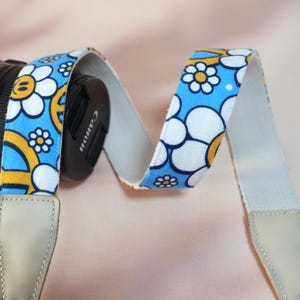 May include: A camera strap with a blue background and a white daisy and yellow peace sign pattern. The strap is attached to a black Canon camera lens. The ends of the strap are light gray.