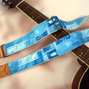 Może przedstawiać: Niebieski pasek do gitary z dżinsu z patchworkowym wzorem. Pasek ma brązowe skórzane końce i metalową klamrę. Pasek jest pokazany na brązowej gitarze akustycznej.
