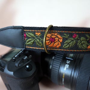 May include: Black camera strap with embroidered floral design. The strap features a black leather shoulder pad and a decorative band with orange, pink, and green flowers. A brass ring connects the strap to the camera.