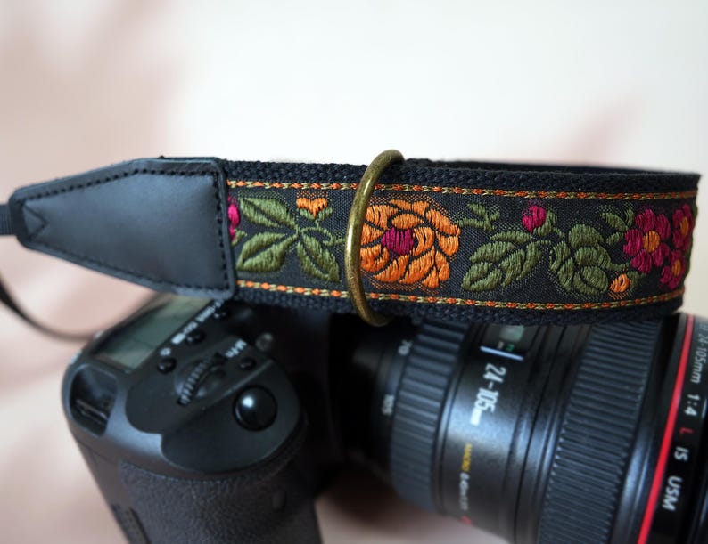 May include: Black camera strap with embroidered floral design. The strap features a black leather shoulder pad and a decorative band with orange, pink, and green flowers. A brass ring connects the strap to the camera.
