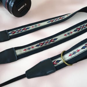 May include: Black camera strap with a woven central band featuring a geometric pattern in red, blue, and white. The strap has black leather ends and a brass-colored metal ring. A Canon lens is visible in the background.