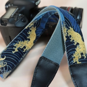 Peut inclure: Une sangle d'appareil photo avec un motif de vagues bleu et or. La sangle a une section centrale grise et des extrémités en cuir noir. Un appareil photo est visible en arrière-plan.