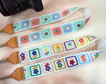 May include: Three colorful camera straps with floral patterns. The straps are white with embroidered squares in various colors, including pink, blue, green, and orange. The ends are tan leather.