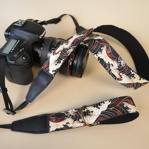 Pode incluir: Uma câmara DSLR Canon preta com lente, apresentando uma correia com um padrão de ondas japonesas em preto, branco e vermelho. A correia inclui pontas em imitação de pele preta e um anel metálico cor de latão para ajuste.