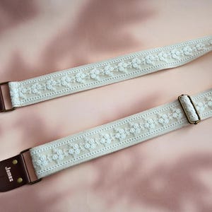 May include: A white guitar strap with a floral design and brown leather ends. The strap has the word "James" embossed on one end.