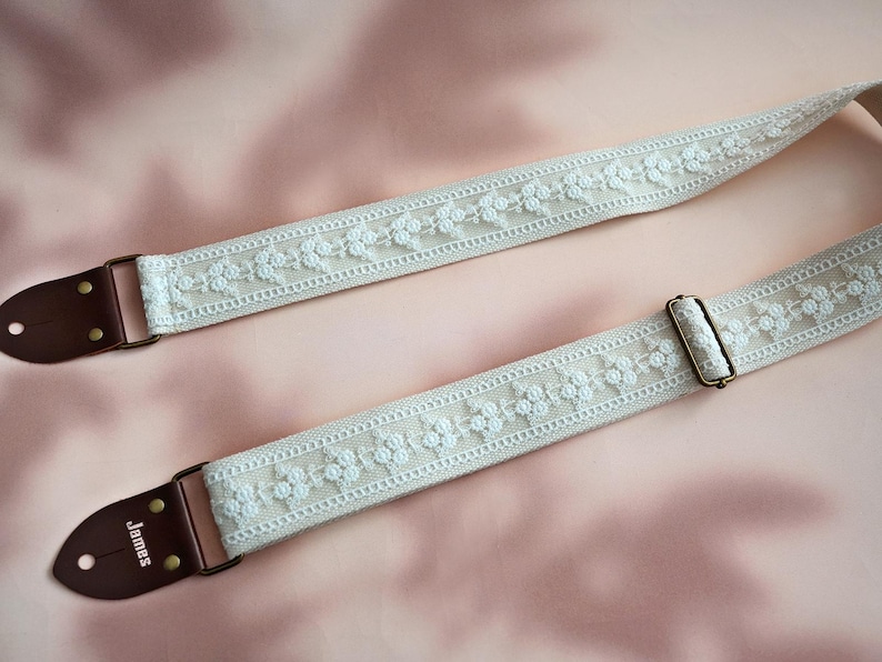 May include: A white guitar strap with a floral design and brown leather ends. The strap has the word "James" embossed on one end.