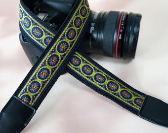 May include: A black camera strap with a decorative pattern in yellow, blue, and orange. The strap features a repeating floral design and black leather ends. The strap is designed to be used with a camera.