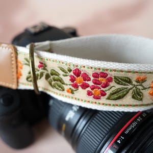 May include: A camera strap with a floral embroidered design. The strap features a cream-colored base with a tan floral pattern in red, orange, and green. The strap has a tan leather end and a metal ring.