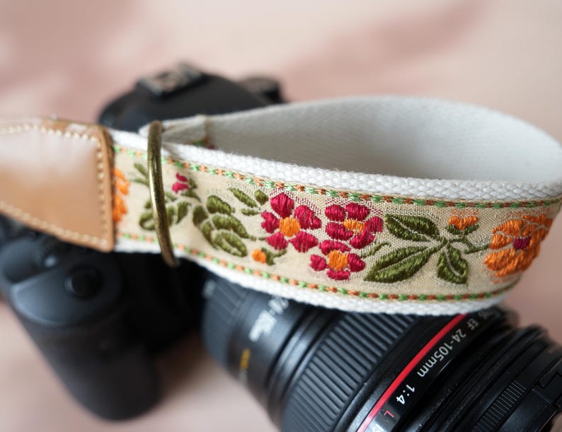 May include: A camera strap with a floral embroidered design. The strap features a cream-colored base with a tan floral pattern in red, orange, and green. The strap has a tan leather end and a metal ring.