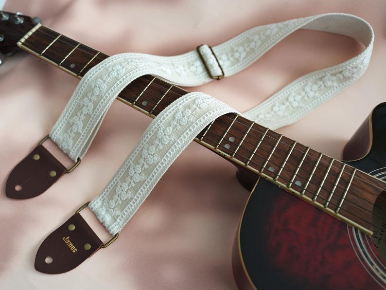 May include: White guitar strap with floral embroidery and brown leather ends. The strap is attached to an acoustic guitar with a red finish.