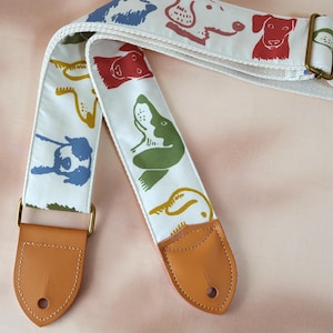 Puede incluir: Correa de guitarra blanca con ilustraciones de perros coloridos en rojo, verde, azul y amarillo. La correa tiene extremos de cuero marr&oacute;n y herrajes met&aacute;licos dorados. La correa mide aproximadamente 5 cm de ancho.