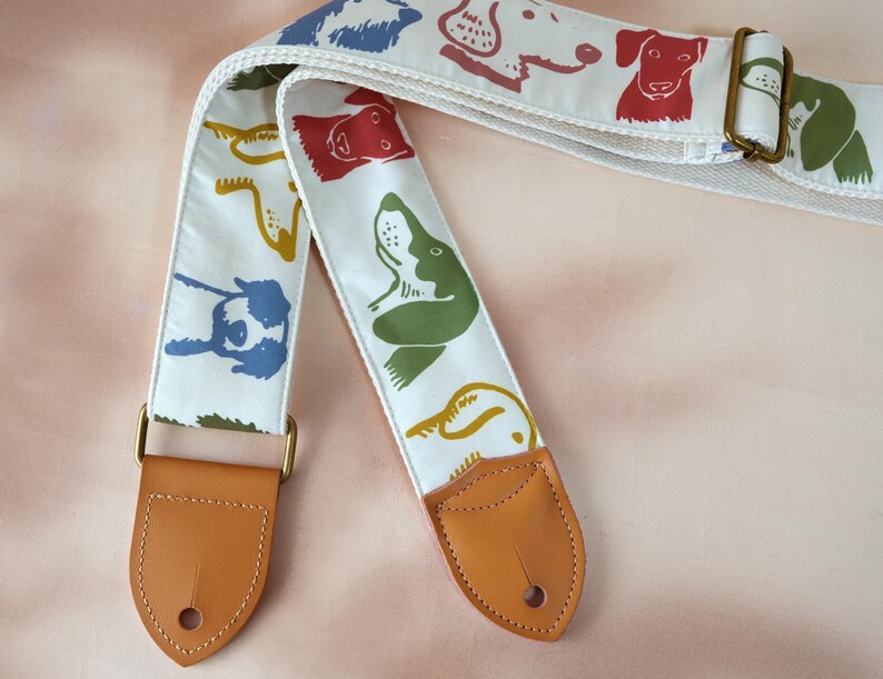 Puede incluir: Correa de guitarra blanca con ilustraciones de perros coloridos en rojo, verde, azul y amarillo. La correa tiene extremos de cuero marr&oacute;n y herrajes met&aacute;licos dorados. La correa mide aproximadamente 5 cm de ancho.