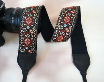 May include: A decorative camera strap with a black background and a repeating pattern of red, white, and blue floral and geometric designs. The strap is attached to a black camera lens.