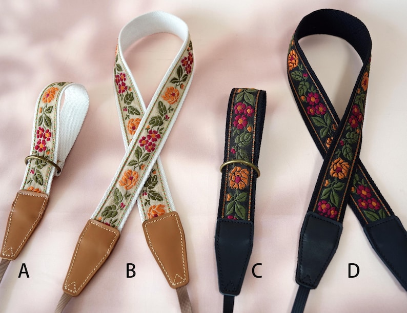 May include: Four camera straps with floral embroidery. Straps A and B have a cream background with orange and red flowers. Straps C and D have a black background with orange and red flowers. All straps have leather ends and metal rings.