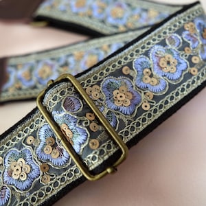 May include: A guitar strap with a floral design. The strap features blue embroidered flowers with gold sequins on a dark gray background. It has a brass-colored metal buckle and brown leather ends.