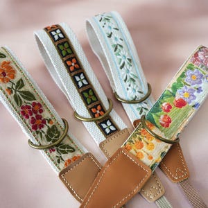 May include: Four decorative camera straps with floral patterns. The straps feature various designs, including orange and red flowers, green leaves, and white backgrounds. Each strap has a brass ring and brown leather ends.