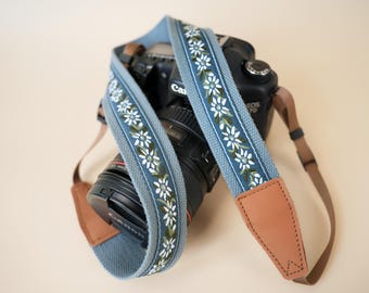 May include: A camera strap featuring a light blue fabric base with an embroidered pattern of white daisies and green leaves. The strap includes tan leather end tabs and black adjustable webbing, shown wrapped around a black digital camera body.