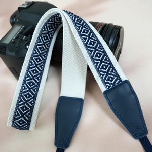 May include: A camera strap with a white center and navy blue geometric patterned edges. The strap has navy blue leather ends and is draped over a black DSLR camera.