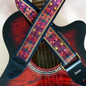 May include: A red acoustic guitar with a black and gold patterned guitar strap. The strap has the word "James" on the end.