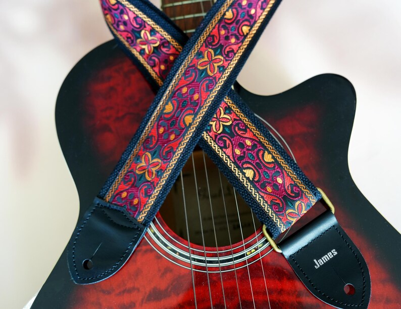 May include: A red acoustic guitar with a black and gold patterned guitar strap. The strap has the word "James" on the end.