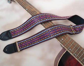 Pode incluir: Uma correia de guitarra colorida com um padrão tecido em roxo, amarelo, azul e vermelho. A correia tem extremidades em couro preto e uma fivela cor de latão. A correia é mostrada sobre uma guitarra.
