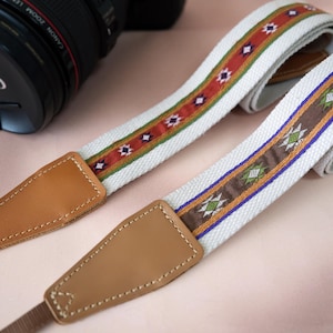 May include: Two camera straps with a white base and colorful woven patterns. One strap features a red and orange design, while the other has a brown and blue pattern. Both straps have tan leather ends.