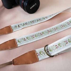 May include: A camera strap with a floral design. The strap is white with a light blue floral pattern in green and pink. The ends are brown leather with a metal ring. A Canon camera is in the background.