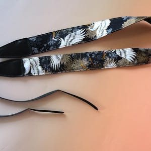 Pode incluir: Correia de câmera preta com um padrão de guindaste branco e dourado. A correia tem duas correias ajustáveis pretas.