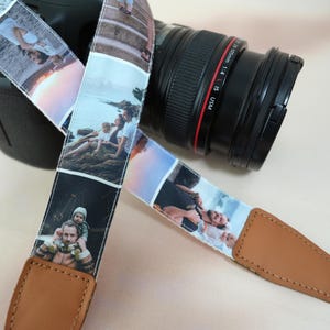May include: A camera strap featuring a collage of family photos. The strap is white with a repeating pattern of images, and has brown leather ends. A black camera with a lens is in the background.