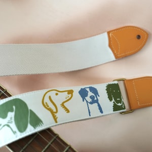 Puede incluir: Correa de guitarra blanca con un patr&oacute;n repetido de ilustraciones de perros en verde, amarillo y azul. La correa tiene extremos de cuero marr&oacute;n y una hebilla de metal dorado. El m&aacute;stil de madera de la guitarra es visible.