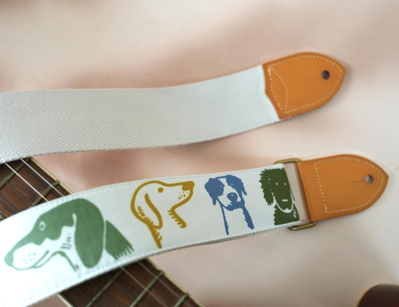 Puede incluir: Correa de guitarra blanca con un patr&oacute;n repetido de ilustraciones de perros en verde, amarillo y azul. La correa tiene extremos de cuero marr&oacute;n y una hebilla de metal dorado. El m&aacute;stil de madera de la guitarra es visible.