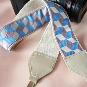 May include: A camera strap with a geometric cube pattern in blue, purple, and white. The strap has a beige fabric base and leather ends. The strap is approximately 30 inches long.