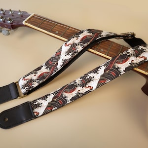 May include: A guitar strap featuring a black, white, and red Japanese-style wave pattern. The strap has black leather ends with circular holes for attachment and is draped over the neck and body of a wooden acoustic guitar.