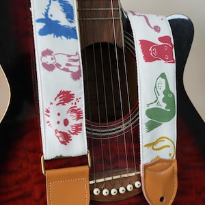 Puede incluir: Correa de guitarra blanca con ilustraciones de perros coloridos en azul, rojo, verde y amarillo. La correa tiene extremos de cuero marr&oacute;n y herrajes met&aacute;licos dorados. La correa est&aacute; sujeta a una guitarra ac&uacute;stica roja y negra.