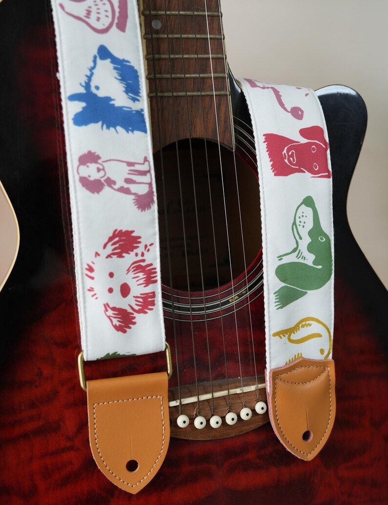 Puede incluir: Correa de guitarra blanca con ilustraciones de perros coloridos en azul, rojo, verde y amarillo. La correa tiene extremos de cuero marr&oacute;n y herrajes met&aacute;licos dorados. La correa est&aacute; sujeta a una guitarra ac&uacute;stica roja y negra.
