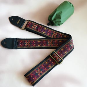 May include: A black guitar strap with a red, gold, and green patterned design. The strap has a leather end and a metal buckle. The brand name "Levys" is printed on the strap.