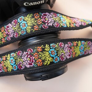 Peut inclure: Sangle d'appareil photo noire avec broderie florale colorée. Le motif répété comprend des fleurs et des feuilles roses, oranges, bleues et vertes. Le mot "Canon" est visible sur le boîtier de l'appareil.