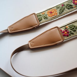 May include: A camera strap with a floral embroidered design. The strap features a tan leather end piece and a woven strap. The embroidered section has orange and pink flowers with green leaves on a light green background.
