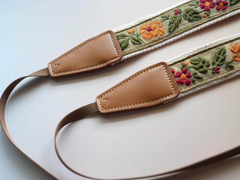 May include: A camera strap with a floral embroidered design. The strap features a tan leather end piece and a woven strap. The embroidered section has orange and pink flowers with green leaves on a light green background.