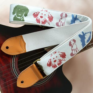 Puede incluir: Correa de guitarra blanca con ilustraciones de perros coloridos en verde, rojo y azul. La correa tiene extremos de cuero marr&oacute;n y herrajes de lat&oacute;n, apoyada sobre una guitarra ac&uacute;stica roja y negra.
