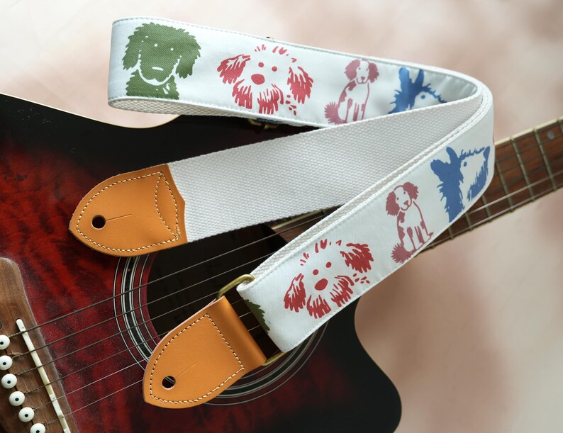 Puede incluir: Correa de guitarra blanca con ilustraciones de perros coloridos en verde, rojo y azul. La correa tiene extremos de cuero marr&oacute;n y herrajes de lat&oacute;n, apoyada sobre una guitarra ac&uacute;stica roja y negra.