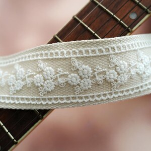 May include: A beige guitar strap with white embroidered floral details. The strap has a decorative buckle.