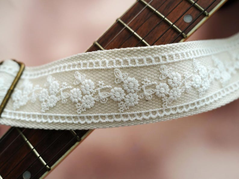 May include: A beige guitar strap with white embroidered floral details. The strap has a decorative buckle.