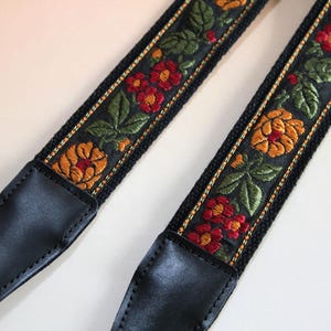 May include: Black camera strap with embroidered floral design. The strap features red, orange, and green flowers and leaves on a black background. The ends are black leather.