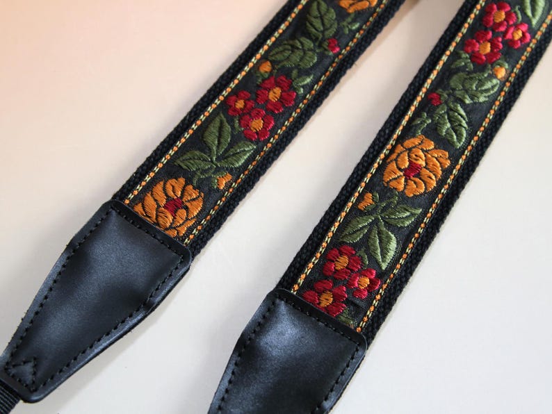 May include: Black camera strap with embroidered floral design. The strap features red, orange, and green flowers and leaves on a black background. The ends are black leather.