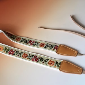 May include: A camera strap with a floral design. The strap features a white base with a woven pattern of orange, red, and green flowers. The ends are tan leather with a tan strap.