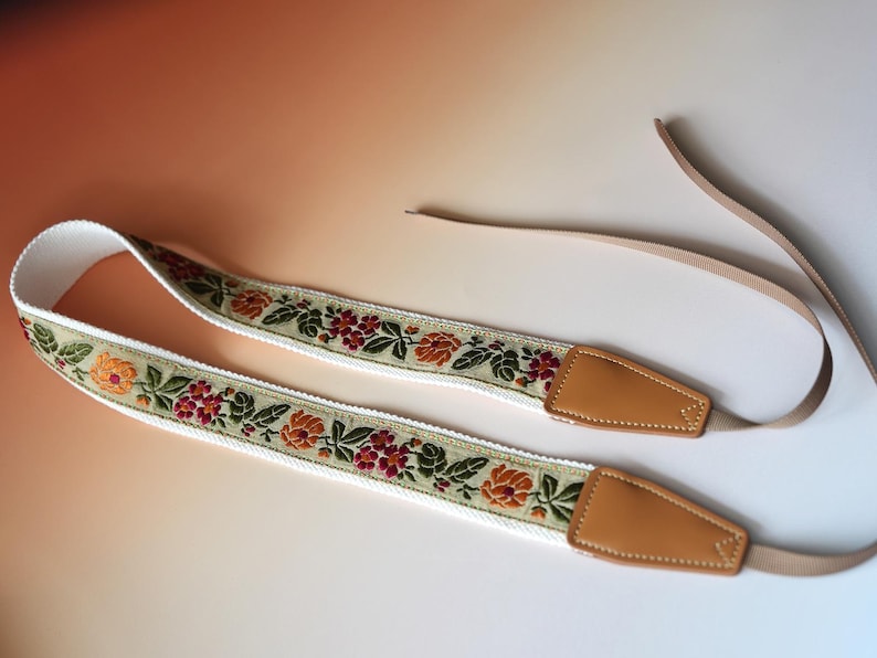 May include: A camera strap with a floral design. The strap features a white base with a woven pattern of orange, red, and green flowers. The ends are tan leather with a tan strap.
