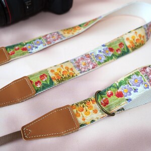 May include: A floral patterned camera strap with brown leather ends. The strap features a repeating pattern of colorful flowers on a light background. The strap is designed to attach to a camera for easy carrying.