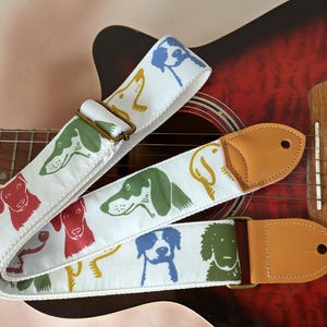 Puede incluir: Correa de guitarra blanca con ilustraciones de perros coloridos en rojo, verde, amarillo y azul. La correa tiene extremos de cuero marr&oacute;n y una hebilla de metal. La correa est&aacute; sobre una guitarra ac&uacute;stica roja.