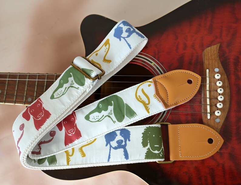 Puede incluir: Correa de guitarra blanca con ilustraciones de perros coloridos en rojo, verde, amarillo y azul. La correa tiene extremos de cuero marr&oacute;n y una hebilla de metal. La correa est&aacute; sobre una guitarra ac&uacute;stica roja.